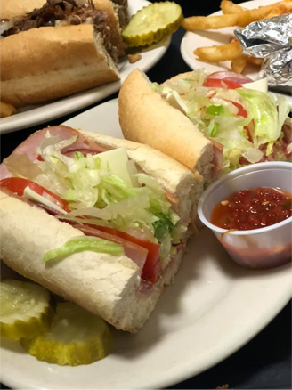 A plate with an italian sub cut in half with pickle slices and a side cup of pepper jelly.
